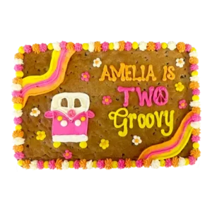 Groovy themed custom cookie cake decorated with colorful buttercream flowers, peace sign, rainbow stripes, and a pink hippie van design, with the message ‘Amelia is Two Groovy.