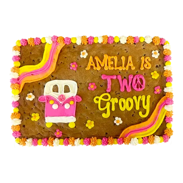 Groovy themed custom cookie cake decorated with colorful buttercream flowers, peace sign, rainbow stripes, and a pink hippie van design, with the message ‘Amelia is Two Groovy.