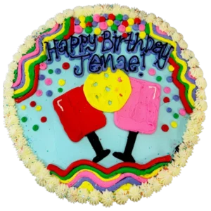 Round cookie cake decorated with colorful frosting that reads “Happy Birthday Jenae!” in blue lettering. The design features two cartoon-style frosted cocktail glasses with, a yellow cookie, and a border of rainbow squiggles, confetti dots, and white star-shaped frosting along the edge.