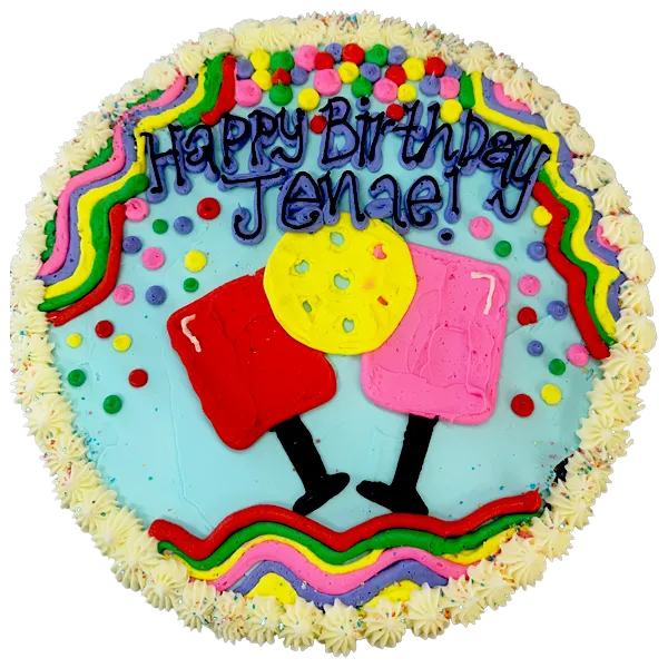 Round cookie cake decorated with colorful frosting that reads “Happy Birthday Jenae!” in blue lettering. The design features two cartoon-style frosted cocktail glasses with, a yellow cookie, and a border of rainbow squiggles, confetti dots, and white star-shaped frosting along the edge.