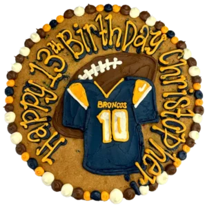 Giant chocolate chip cookie cake, decorated in a football theme with a frosted jersey and football and happy birthday text.