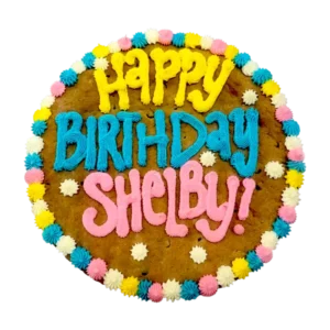 Giant chocolate chip cookie cake, frosted with a bright and colorful border and text that says "happy birthday Shelby".