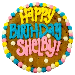 Giant chocolate chip cookie cake, frosted with a bright and colorful border and text that says "happy birthday Shelby".