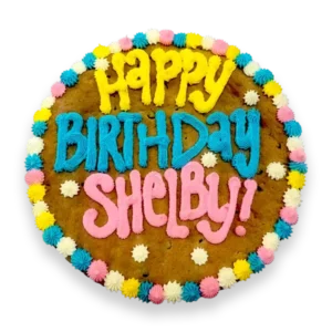 Giant chocolate chip cookie cake, frosted with a bright and colorful border and text that says "happy birthday Shelby".