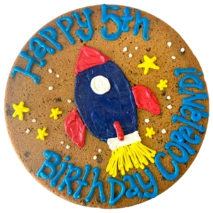 Giant chocolate chip cookie cake decorated with a frosted rocket ship, stars, and text that reads "happy birthday Copeland".