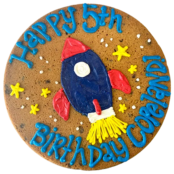 Giant chocolate chip cookie cake decorated with a frosted rocket ship, stars, and text that reads "happy birthday Copeland".